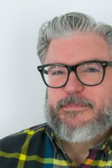 Photo of author Alonso Duralde. The author has salt and pepper hair, mustache, and beard, and wears black glasses. He is looking at the camera in a candid shot.